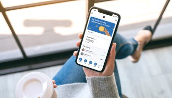 A woman views her cryptocurrency balance on her PayPal app on her phone while holding coffee.