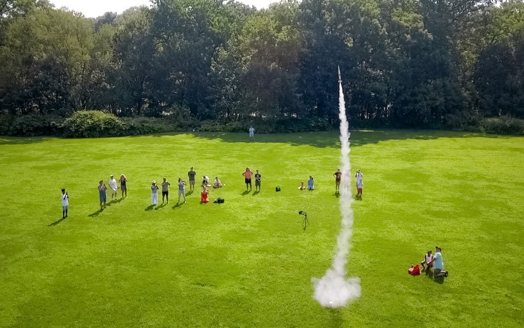 A crowd of people stand in an open field watching a model rocket take off.