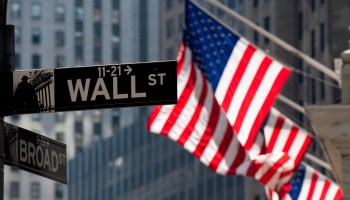 Three American flags fly outside the New York Stock Exchange. In the foreground is a street sign for Wall Street.