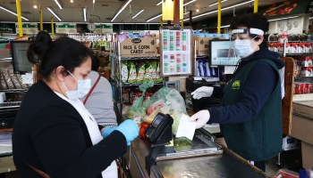 A shopper and cashier both wear masks, gloves and the cashier also has on a plastic visor at the checkout station Pat's Farms grocery store on March 31, 2020 in Merrick, New York.