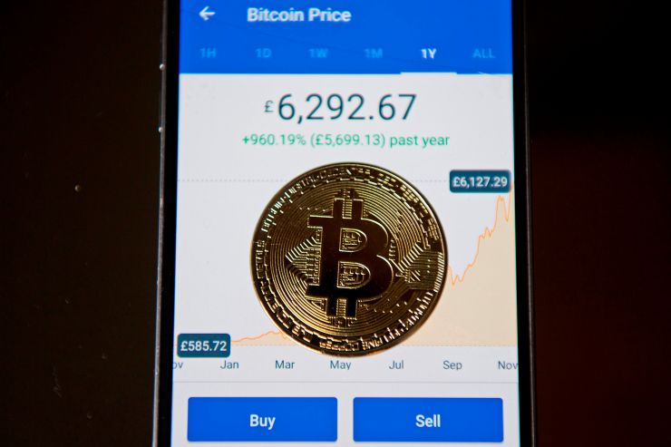 A gold plated souvenir Bitcoin coin is arranged for a photograph on a smart phone displaying current value of a single bitcoin, and options to buy or sell, on an app for the digital asset broker, Coinbase.