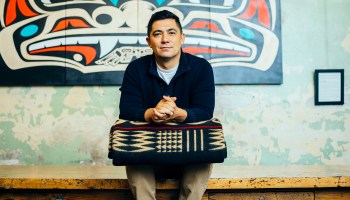 Louie Gong, founder and CEO of Eighth Generation, sits on a bench in front of a piece of Native art holding a wool blanket.