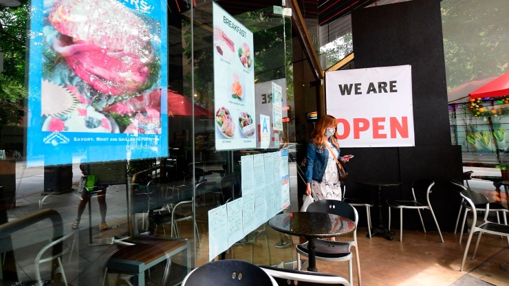 A woman steps out of a restaurant with her takeout order. Behind her is a large "We are open" sign.