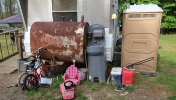 Side view of a silver trailer home with a tan porta-potty on one side and a gray mobilehandwashing station next to it. A rusted oil tank is against the house, and leaned against it are two childrens bicycles and a small plastic pink chair.