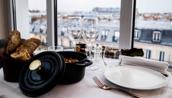 A table is set with glasses, plates, silverware, a basket of bread and a dutch oven. The table is set by the window of a hotel room.