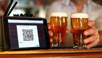 A person holds two beers next to a screen with a QR code pulled up.