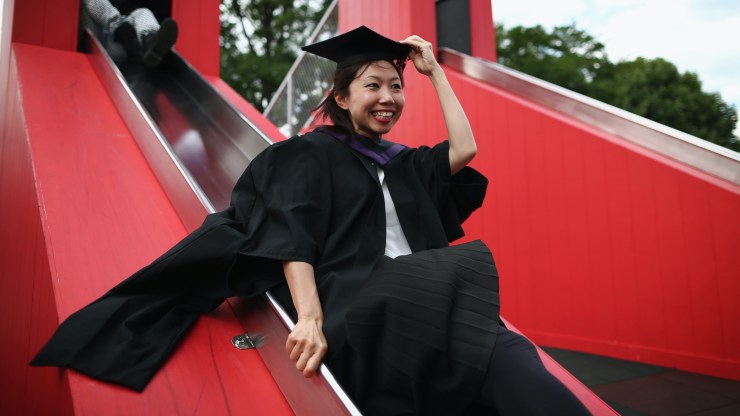 A student in a graduation cap and gown goes down a slide.