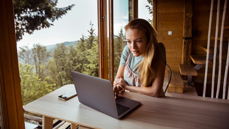 A woman works on a laptop from her cabin in the woods. For high earners who are able to work remotely, the desire for a different lifestyle may prompt a relocation.
