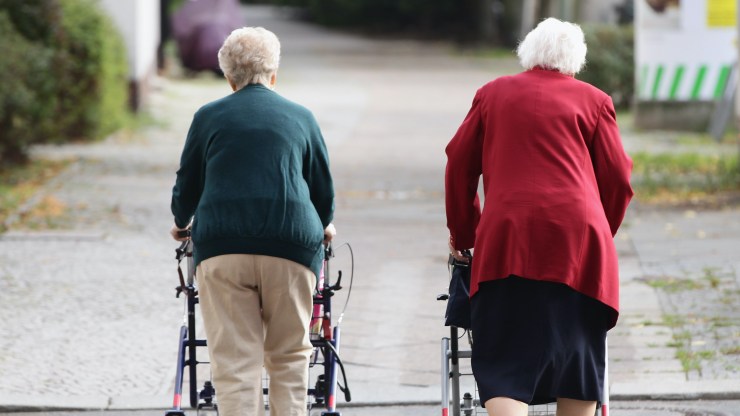 Two elderly women take a stroll with the aid of walkers.