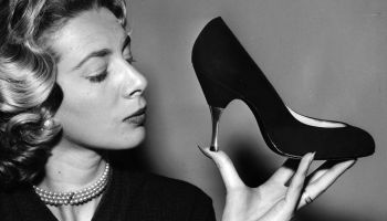 A black and white photo of a woman holding a high heel in the palm of her hand.
