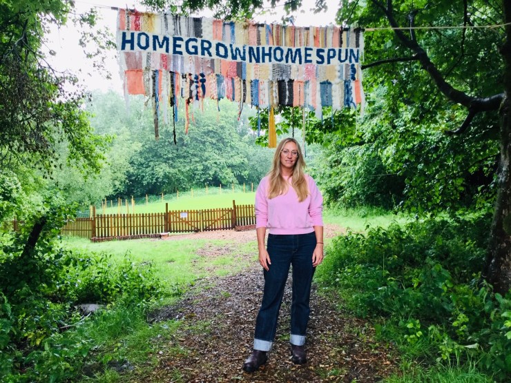 Justine Aldersey-Williams poses in front of a natural path, above which a banner of fabric says "HOMEGROWN. HOMESPUN."