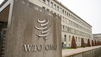 The WTO headquarters in Geneva, Switzerland, photographed at an angle, emphasizes the long, stone-colored building with arched doorways on the ground level and rows of paned windows above.