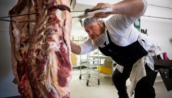 A man with a beard, hairnet and apron saws through a hunk of beef.