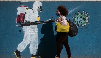 A woman wearing a mask walks past a wall with a graffiti depicting a cleaner in protective gear spraying viruses with the face of President Jair Bolsonaro in Estacio neighborhood amidst the coronavirus (COVID-19) pandemic on June 8, 2020 in Rio de Janeiro, Brazil.