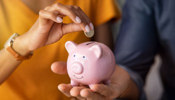 This photo illustration shows someone holding a pink piggy bank while another person puts a coin in it.