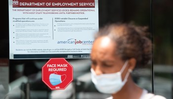 A Black woman wearing a mask walks in front of a Department of Employment Service sign.