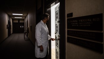 A doctor in a white lab coat and mask opens an office door.