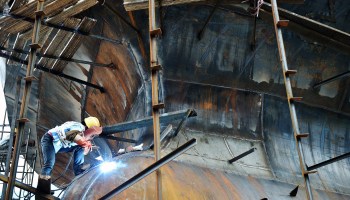 A person welds metal on the underside of a massive shipping vessel.