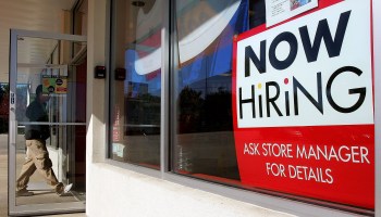 A man enters a store where a "Now Hiring" sign is prominently displayed.