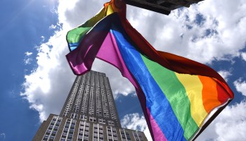 A Pride flag flies with a skyscraper in the background on 5th Avenue in New York City.