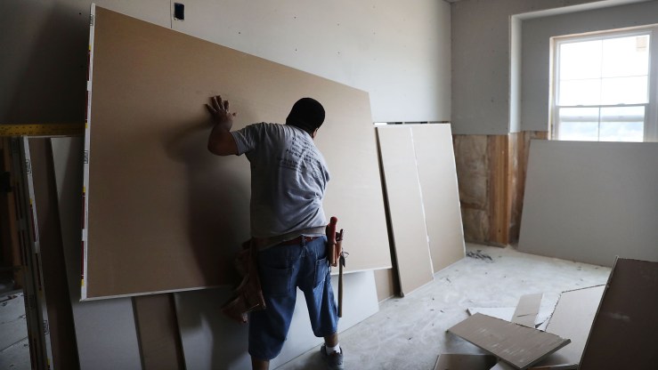 A man lifts a sheet of drywall in a partially renovated room.