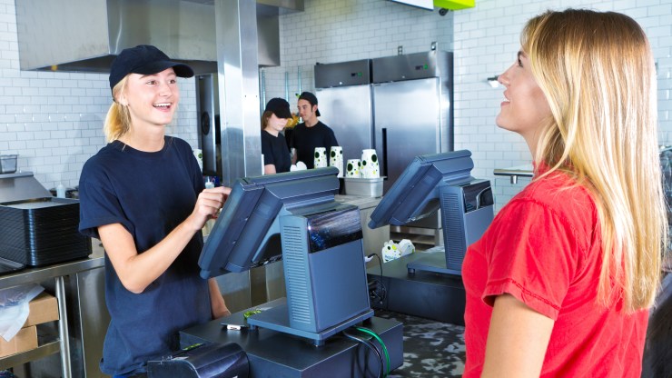 A teenage female server staff is assisting a woman placing her order at the checkout cashier counter with the kitchen staff working in the background.