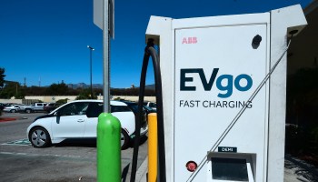 A white car is plugged into a charging station labeled, "EV go Fast Charging," in a large Walmart parking lot.