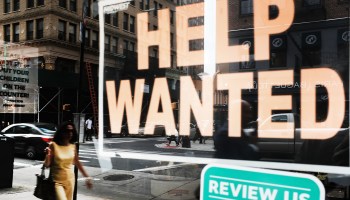 The reflection of a woman can be seen in a window, where a "Help Wanted" sign is prominantly displayed.