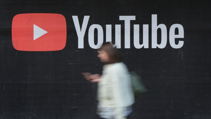 A woman in white walks past a black billboard where the word "YouTube," and its red logo of a play button are inscribed.