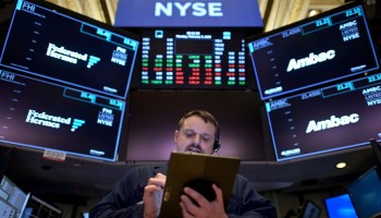 A person with a tablet and headset stands in front of market numbers at the New York Stock Exchange.