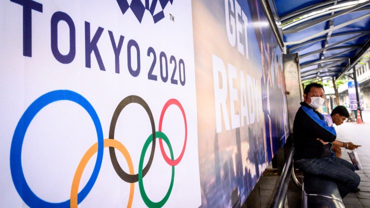 A man at a bus stop bench sits in front of an advertisement for the Olympics that reads "Tokyo 2020. Get ready."