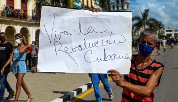In Havana, a man wearing a striped red and black shirt and a face mask holds a sign that says, "Viva la Revolución Cubana"
