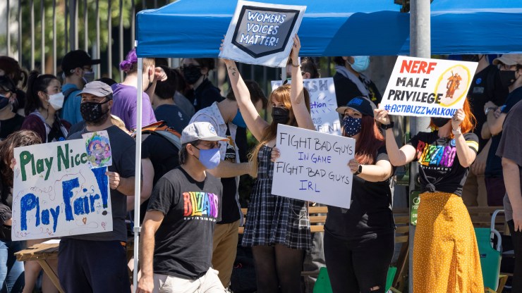 At the Activision Blizzard Walkout, on July 28, a group of four people wearing masks stand in front of a large, masked crowd holding signs. One person, wearing a "Blizzard Entertainment" T-Shirt stands holding a sign that says, "NERF Male Privilege." Another person, wearing a plaid jumpsuit, holds a sign that says, "women's voices matter"
