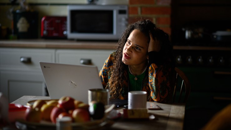 A person runs their hands through their hair, looking stressed, as they sit on their laptop at a kitchen table.