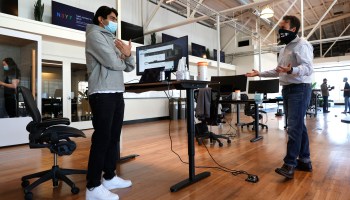 Two men with masks chat at standing desks.