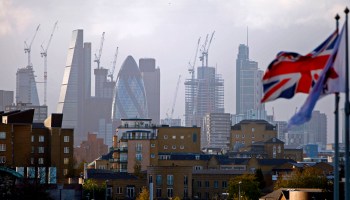The national flag of the U.K. flies in the foreground, and the City of London and its skyscrapers are in the background.