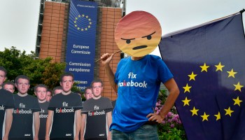 A member of the advocacy group Avaaz, wearing an angry emoji mask, stands in front of the EU headquarters and over a dozen cutouts of Mark Zuckerburg wearing "fix Facebook" shirts.