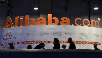 At a las vegas convention, a sign shows "Alibaba.com" in bright orange letters as a group of people walk on a ramp below it,