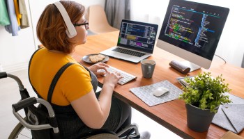 Female freelance programmer in modern headphones sitting in wheelchair and using computers while coding web game at home