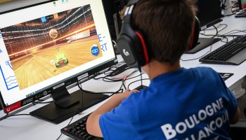 A young boy plays a driving video game on a desktop computer. He is wearing a blue t-shirt and his back is facing the camera.