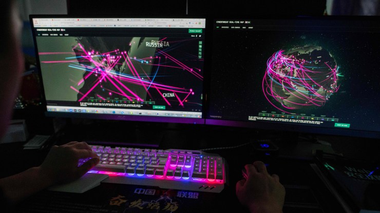 In a dimly lit room, two computer screens show world maps and pink rays to signify different cyberattacks. A pair of hands rests on a blue and pink-lit keyboard as the hacker navigates the desktop.