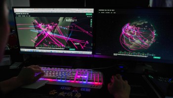 In a dimly lit room, two computer screens show world maps and pink rays to signify different cyberattacks. A pair of hands rests on a blue and pink-lit keyboard as the hacker navigates the desktop.