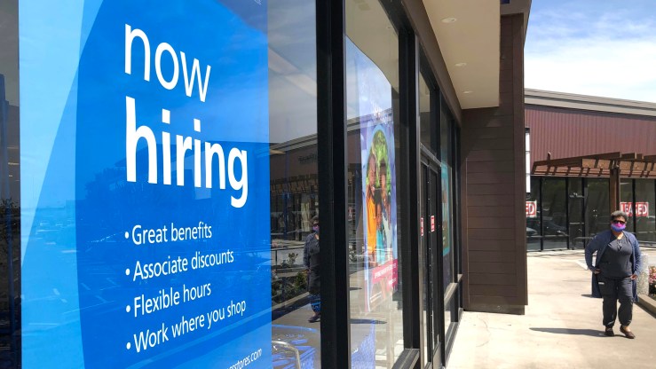 A pedestrian passes a Now Hiring sign on a retail store window.