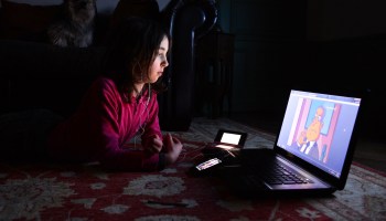 A young girl wearing a pink sweater watches a YouTube cartoon while lying on a red bedspread in a dark room.