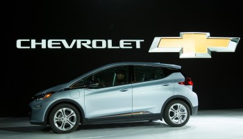 A silver Chevrolet Bolt is featured on a stage with a black background behind it that states, "Chevrolet" and includes the company's signature gold and silver cross.