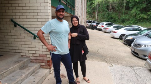 Amal Abuldood, wearing black leggings, a black long-sleeved shirt, hijab, and sandals stands next to her husband, Atheer Abdullah, who is wearing dark jeans, a light blue t-shirt, and a slightly darket blue baseball cap. 