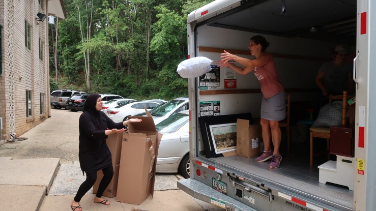 Laura Thompson Osuri, wearing a pink t-shirt with rolled up sleeves and jean shorts, tosses a pillow from the back of an open U-Haul truck. On the ground and ready to catch the pillow is a smiling Amal Abuldood, wearing black pants, a long sleeved black shirt, and a flowy black hijab.