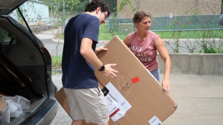 Homes Not Borders executive director Laura Thompson Osuri, , wearing a pink t-shirt with rolled up sleeves, moves a large cardboard box containing a bedframe with the help of her nephew, Jack Campbell, who is wearing a blue t-shirt and khaki shorts. He is tall and has to duck under the open door of the minivan they are pulling the box out of. 