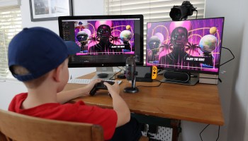 The photographer's son, Ansel, plays Fortnite on two separate computer monitors as he sits at a wooden desk. On the screen is an avatar of Travis Scott as part of his performance on the platform in April 2020.