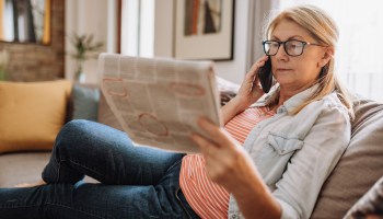 A middle-aged woman views newspaper listings while speaking on the phone.
